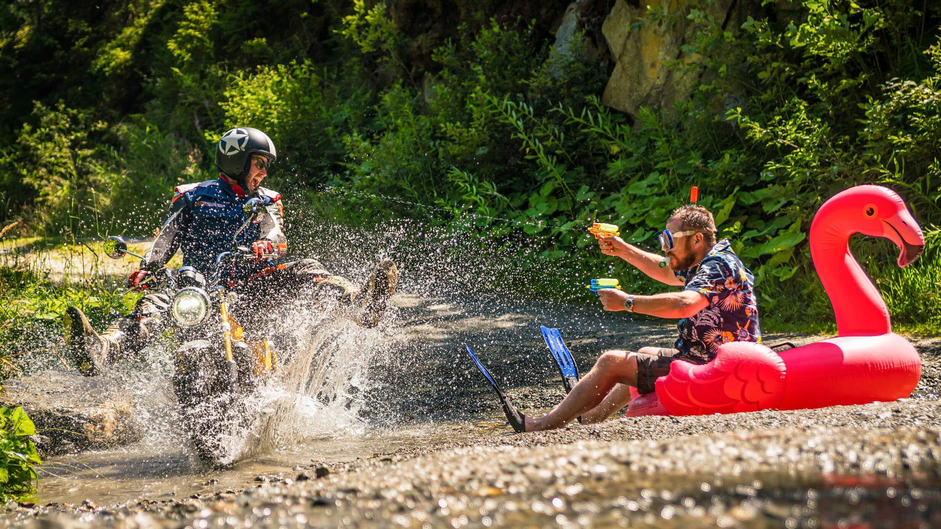Mann auf Motorrad spritzt Wasser auf Mann mit Wasserpistolen im Flamingo-Schwimmring (© Moppetfoto.de)