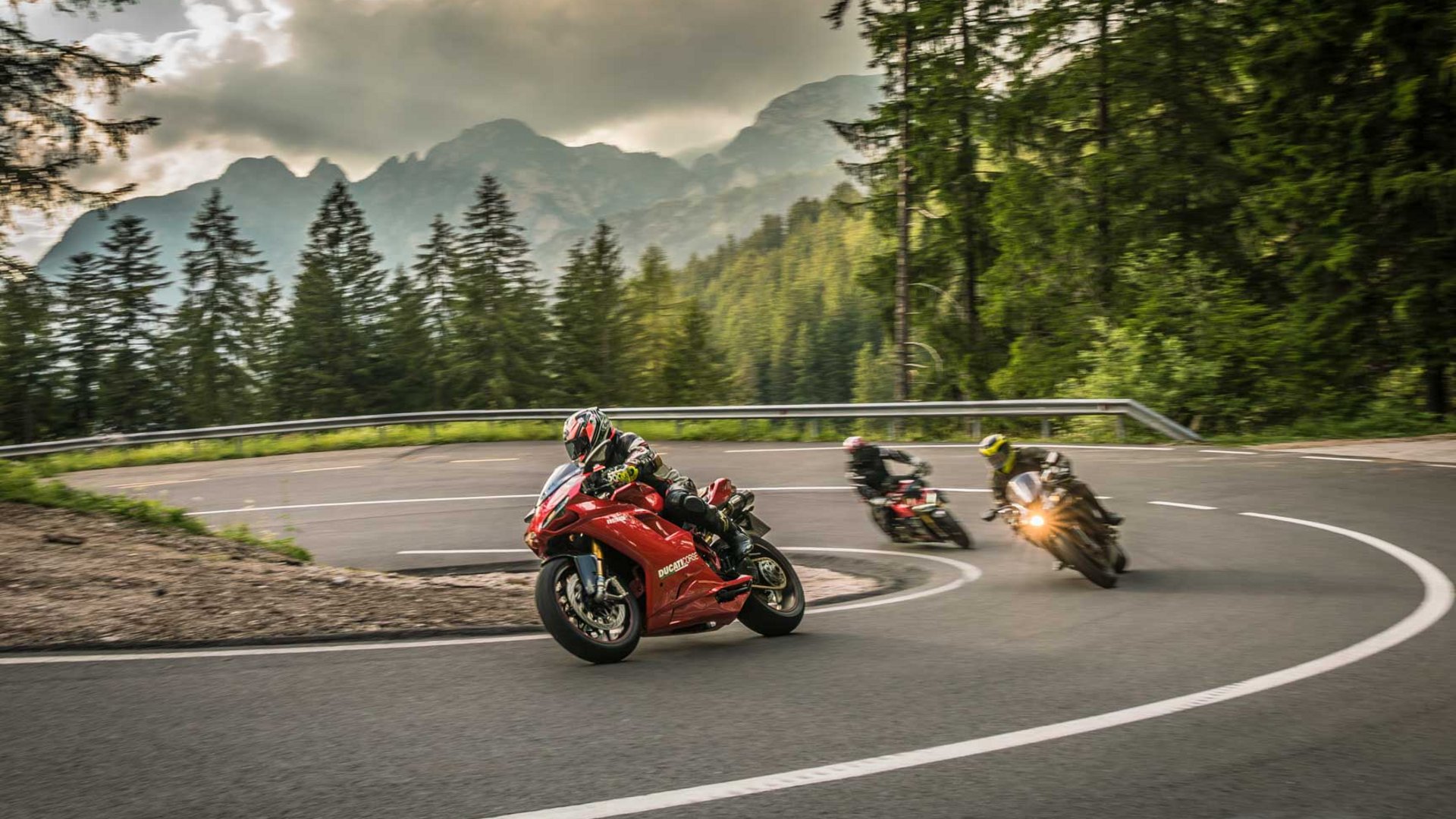 Drei Motorradfahrer fahren eine kurvige Bergstraße mit Wald und Bergen im Hintergrund (© Moppetfoto.de)